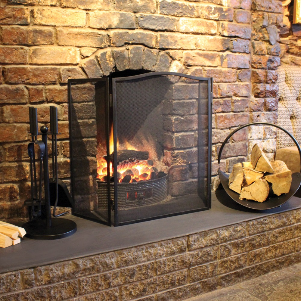 Folding fire screen in front of a lit fireplace with logs and tools on a stone hearth.