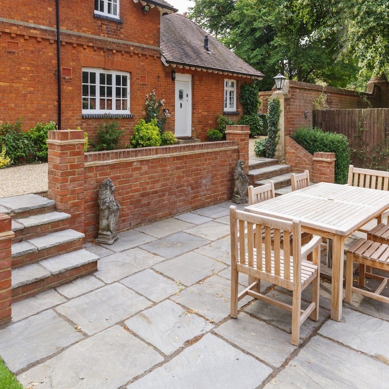A patio area with Indian sandstone paving, wooden outdoor furniture, and a brick house in the background.