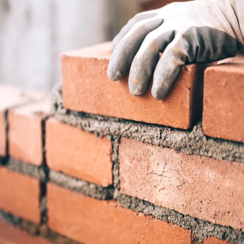 A gloved hand placing a brick on a wall with mortar.