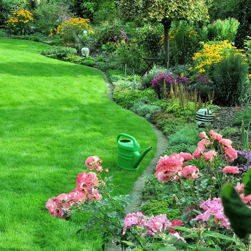 A lush green lawn bordered by vibrant flowers and a green watering can on the path.