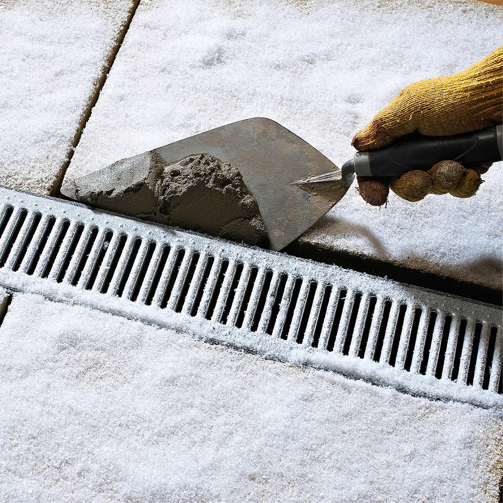 A hand wearing a glove using a trowel to apply mortar on a snowy surface next to a drainage grate.