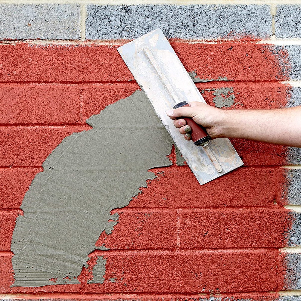 A hand applying grey render to a red brick wall using a trowel.