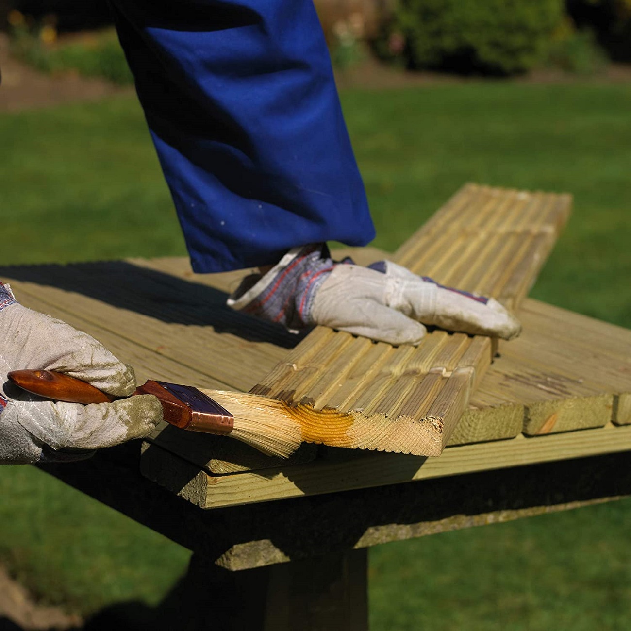 Person applying clear wood preserver to wooden planks outdoors with a brush.
