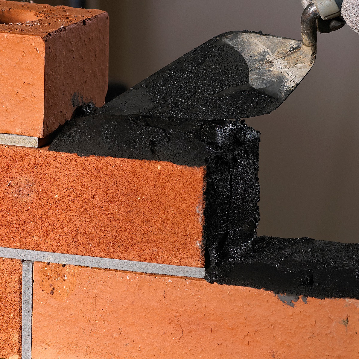 Close-up of black mortar applied between red bricks using a trowel.