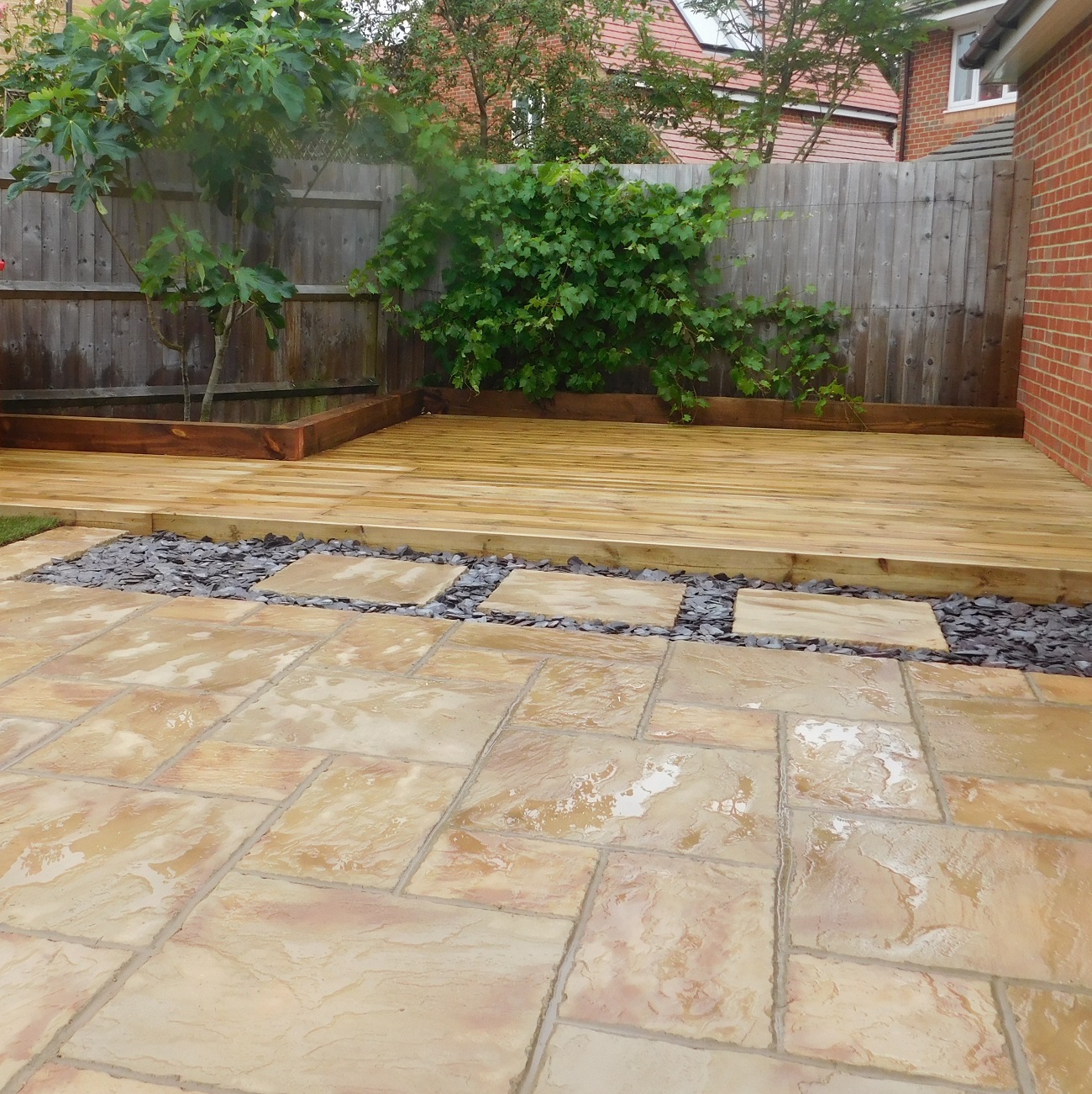 A clean patio with stone tiles and a wooden deck, surrounded by a garden fence and greenery.