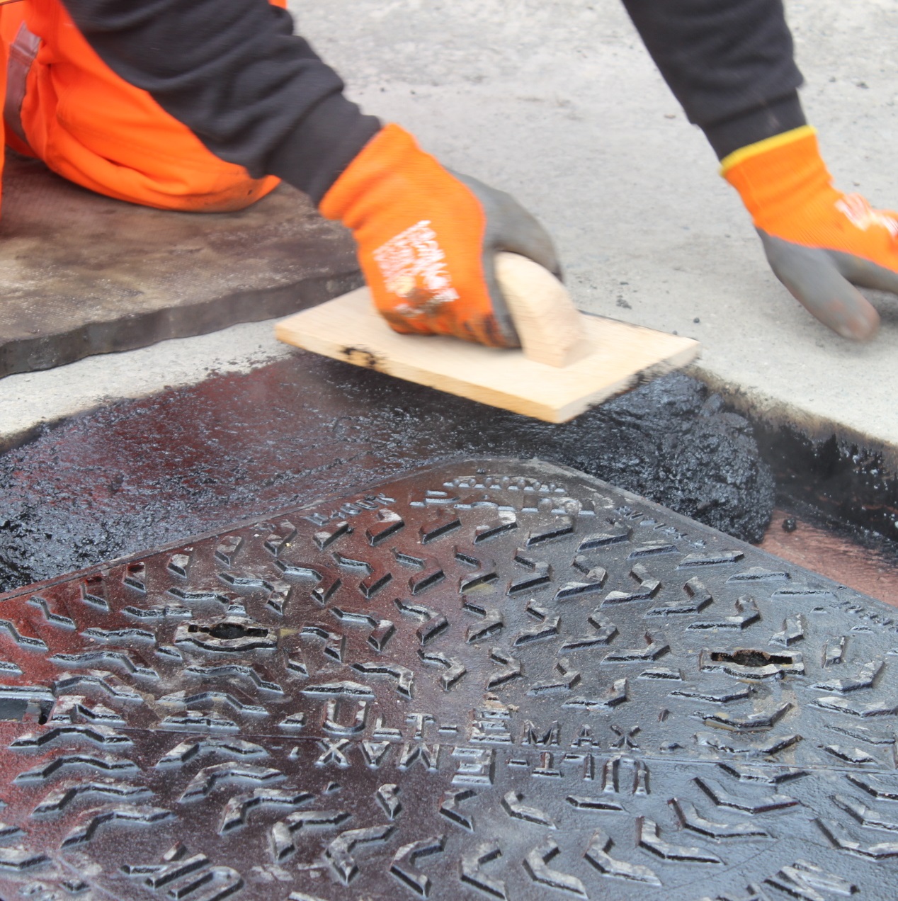 Worker applying Bond It Instant Pot Hole Repair Tarmac on a road surface near a manhole cover.