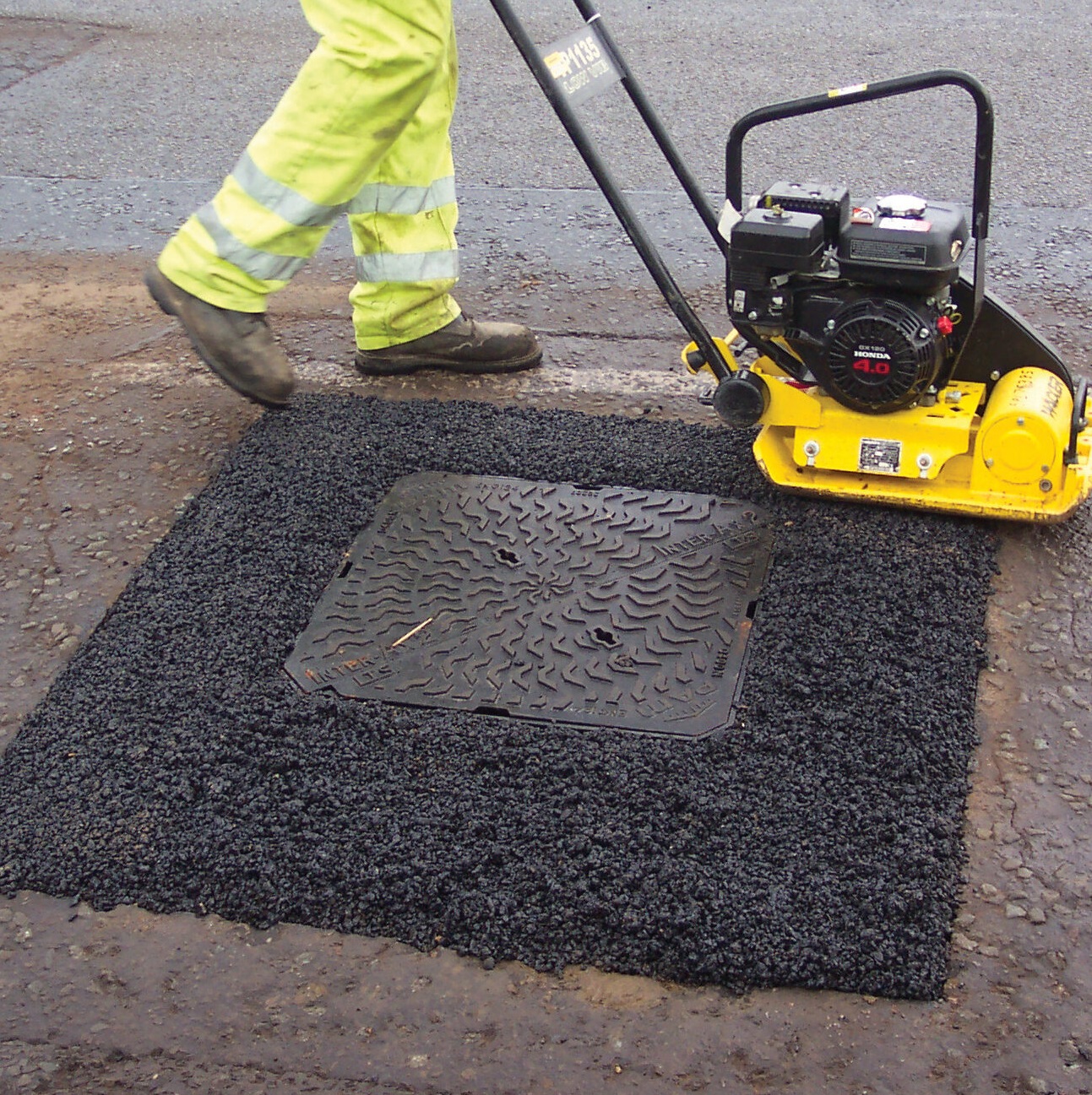 Worker using a compactor on freshly applied tarmac around a manhole cover.