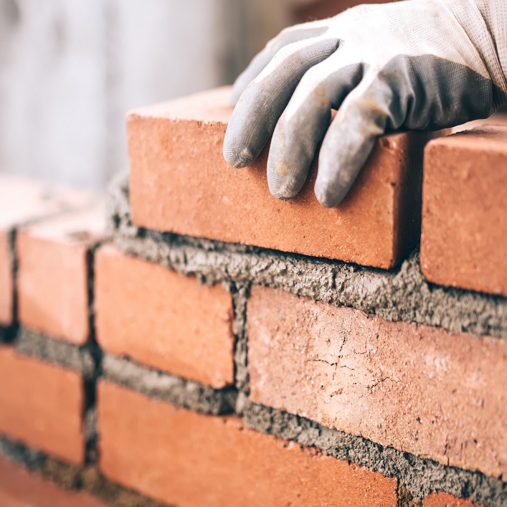A gloved hand placing a brick onto a wall with mortar.