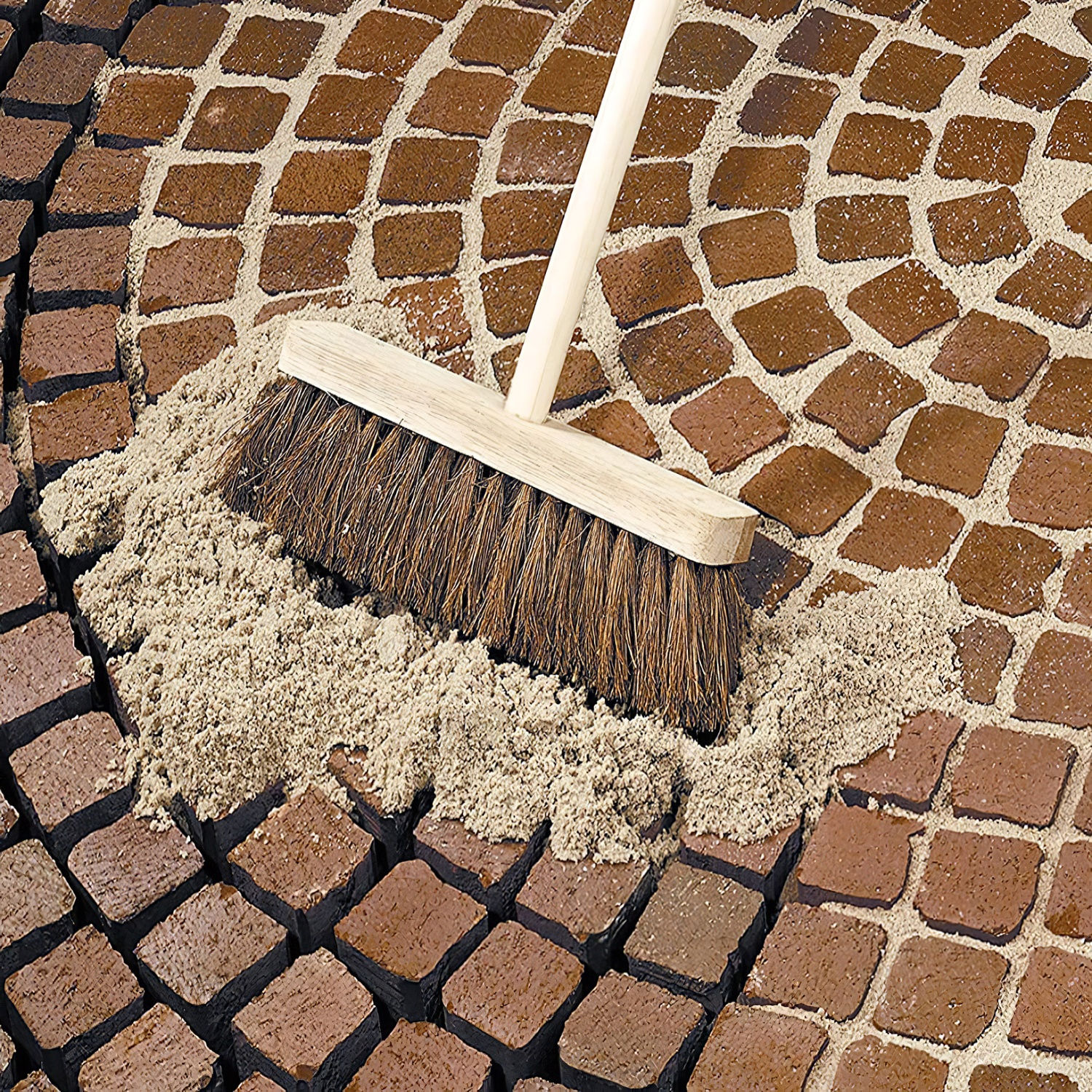 Broom sweeping fine sand into narrow joints of red brick paving