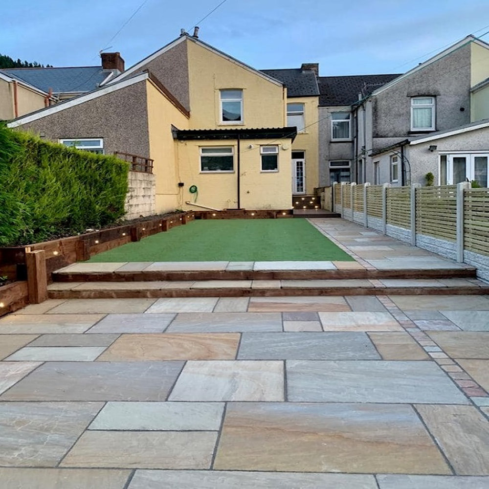Backyard with natural stone paving and artificial grass, bordered by wooden steps and fencing.