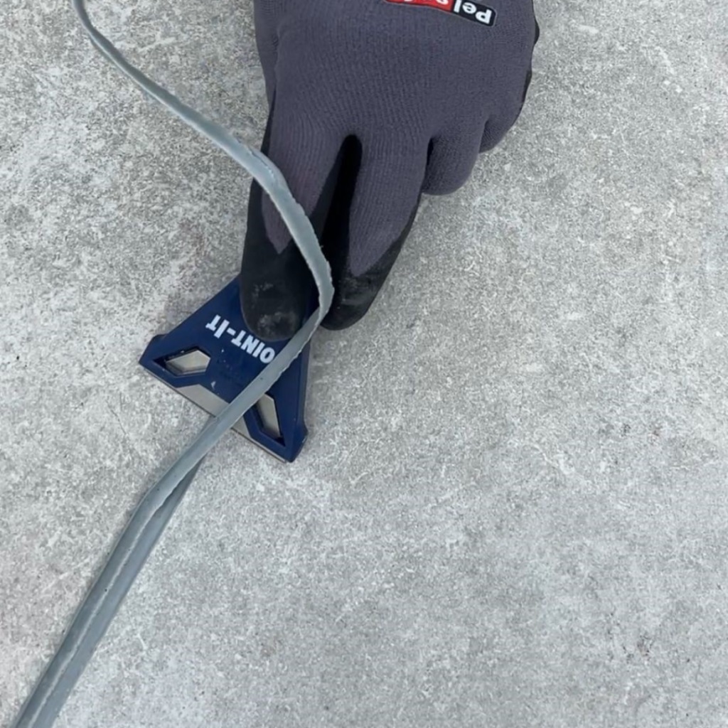 Gloved hand using a Joint It Porcelain Epoxy Grout Removal Scraper on a tiled surface.