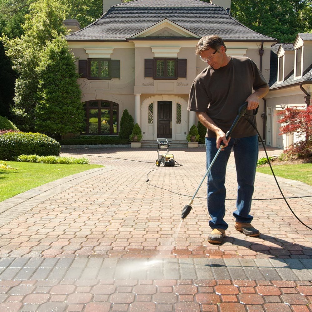 Person using a pressure washer on a brick driveway in front of a house.