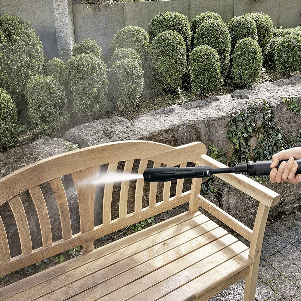 Person using a pressure washer to clean a wooden bench in a garden setting.
