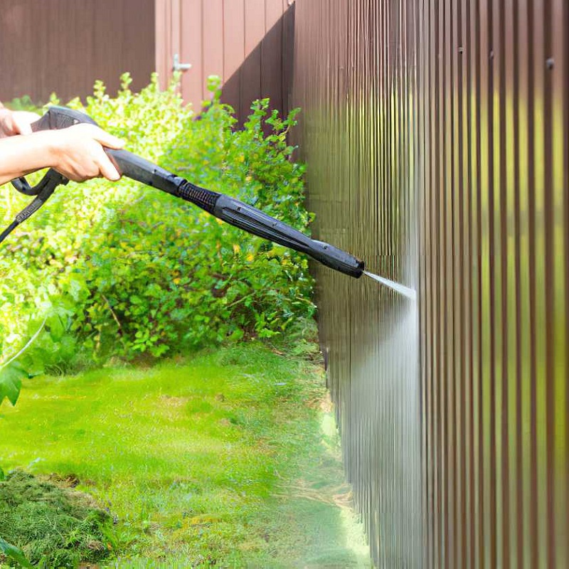Person using a pressure washer to clean a wooden fence in a garden setting.