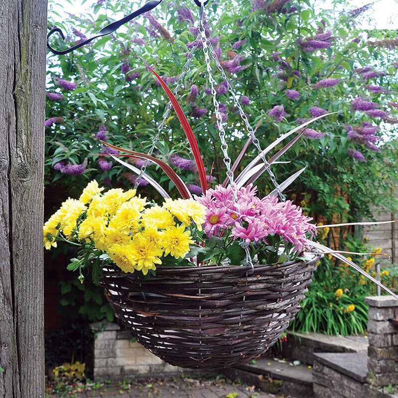 Round hanging flower basket made of dark rattan willow wicker, filled with yellow and pink flowers, hanging in a garden setting.