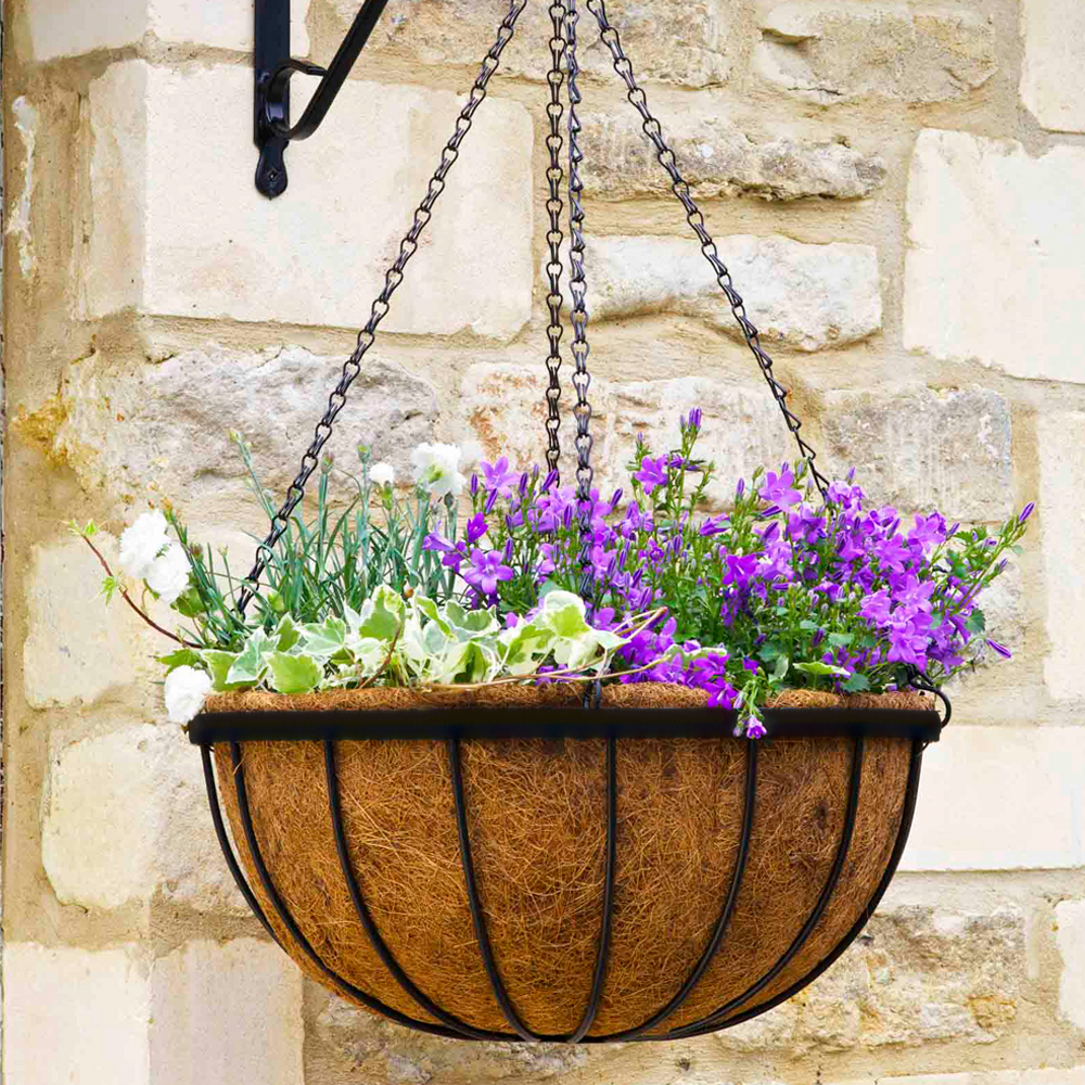 A 12 inch round hanging basket with a coco liner, filled with purple and white flowers, attached to a stone wall.