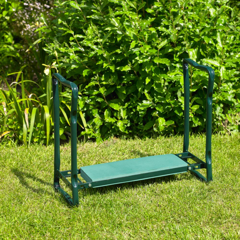 Green folding garden kneeler with handles on grass, surrounded by lush plants.