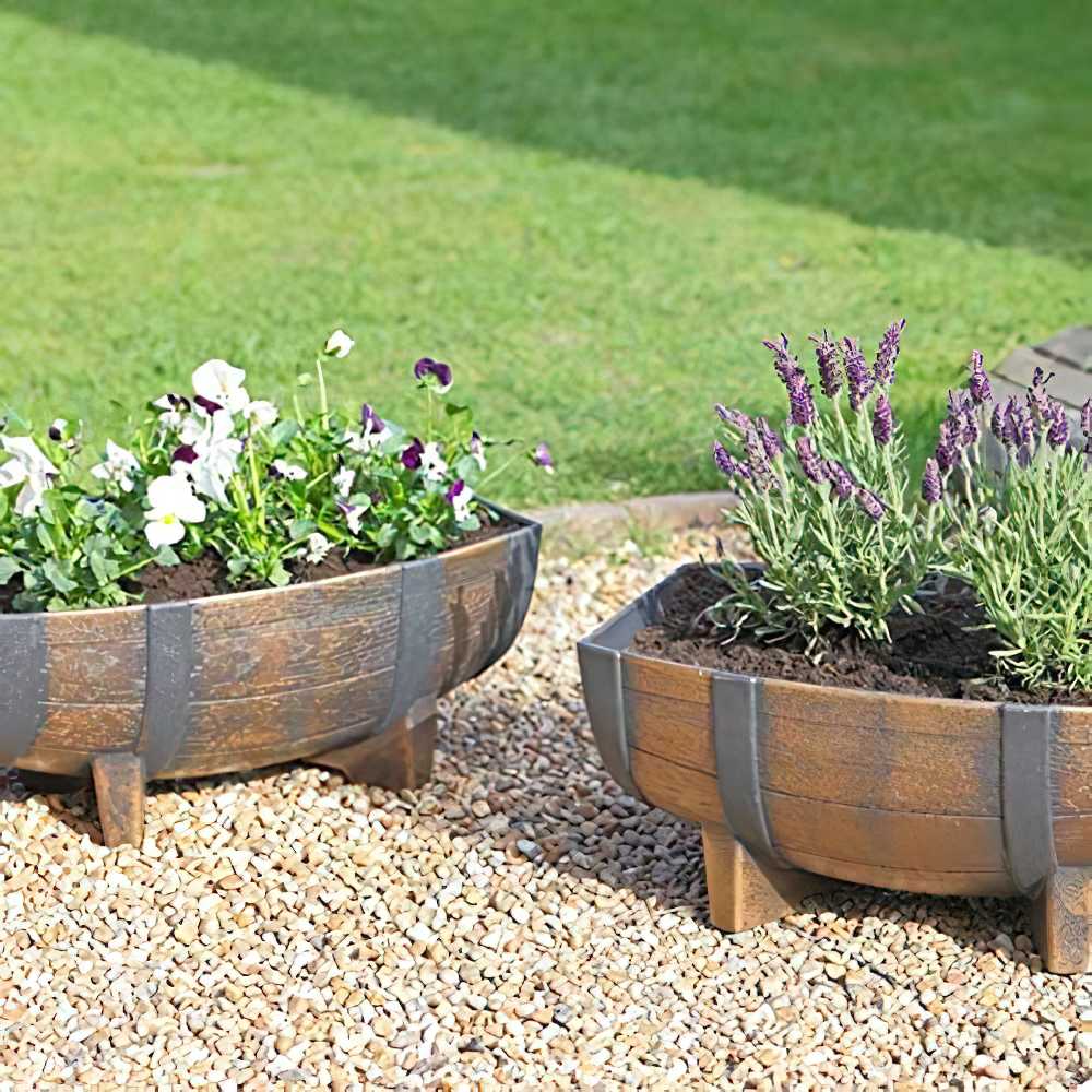Two Kingfisher Garden Wood Effect Half Barrel Plastic Planters with flowers and plants on a gravel surface.