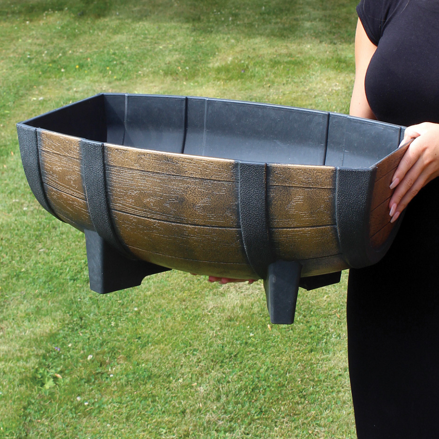 A person holding a Kingfisher Garden Wood Effect Half Barrel Plastic Planter Trough on a grassy lawn.