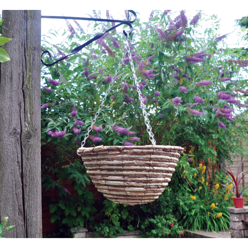 A natural rope 12 inch hanging flower basket suspended from a metal bracket, with lush green foliage and purple flowers in the background.
