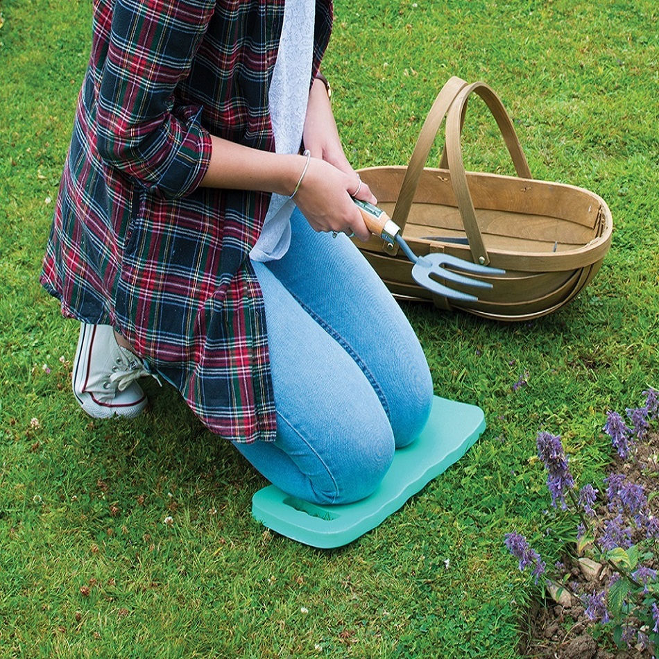 Person kneeling on a green knee protection pad while gardening on grass, with a basket and garden tool nearby.