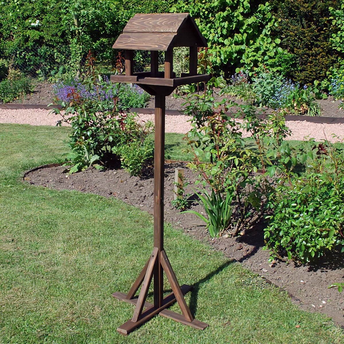 A rustic wooden bird table standing in a garden with greenery and flowers.