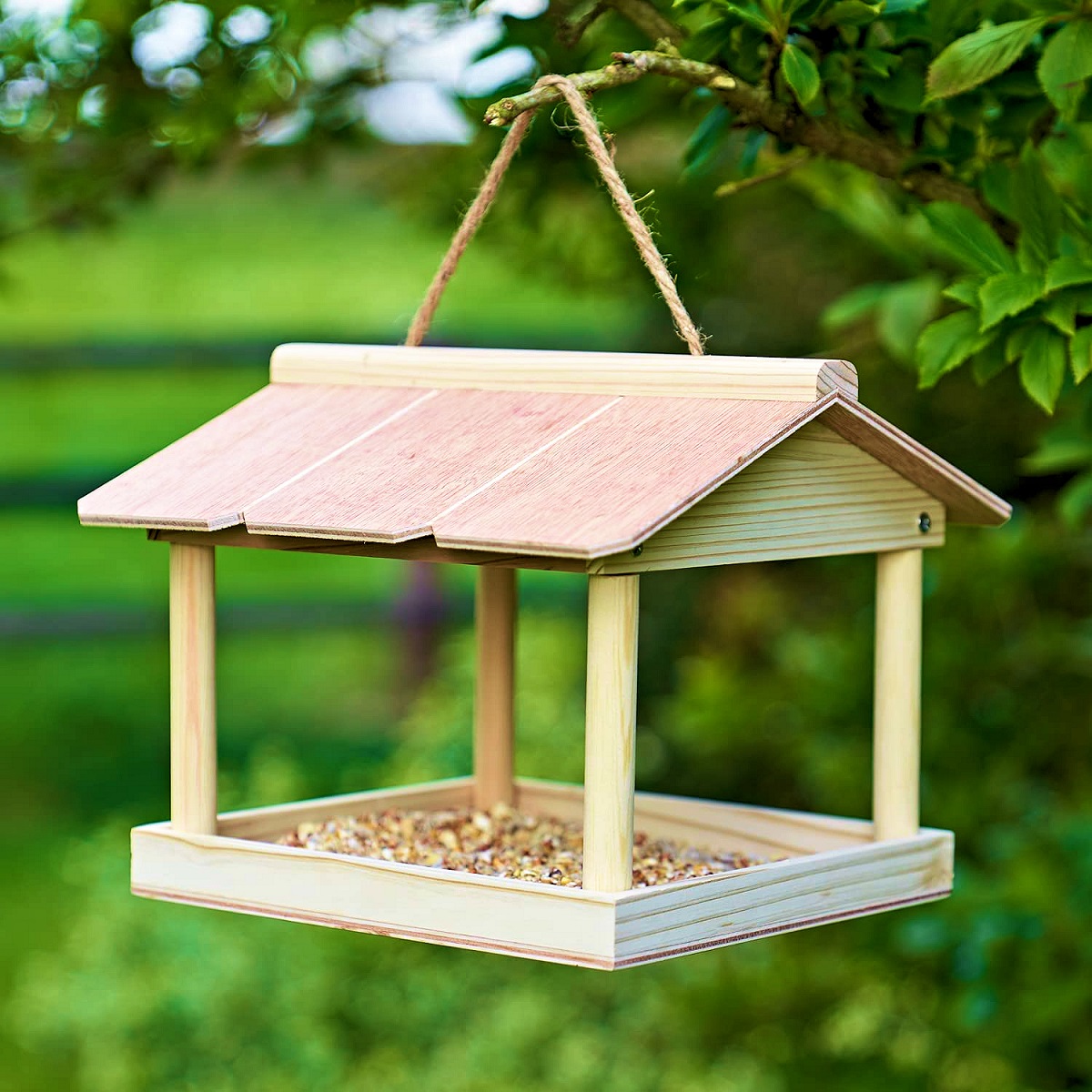 Hanging wooden bird feeder table with a roof, filled with seeds, suspended from a tree branch in a garden setting.