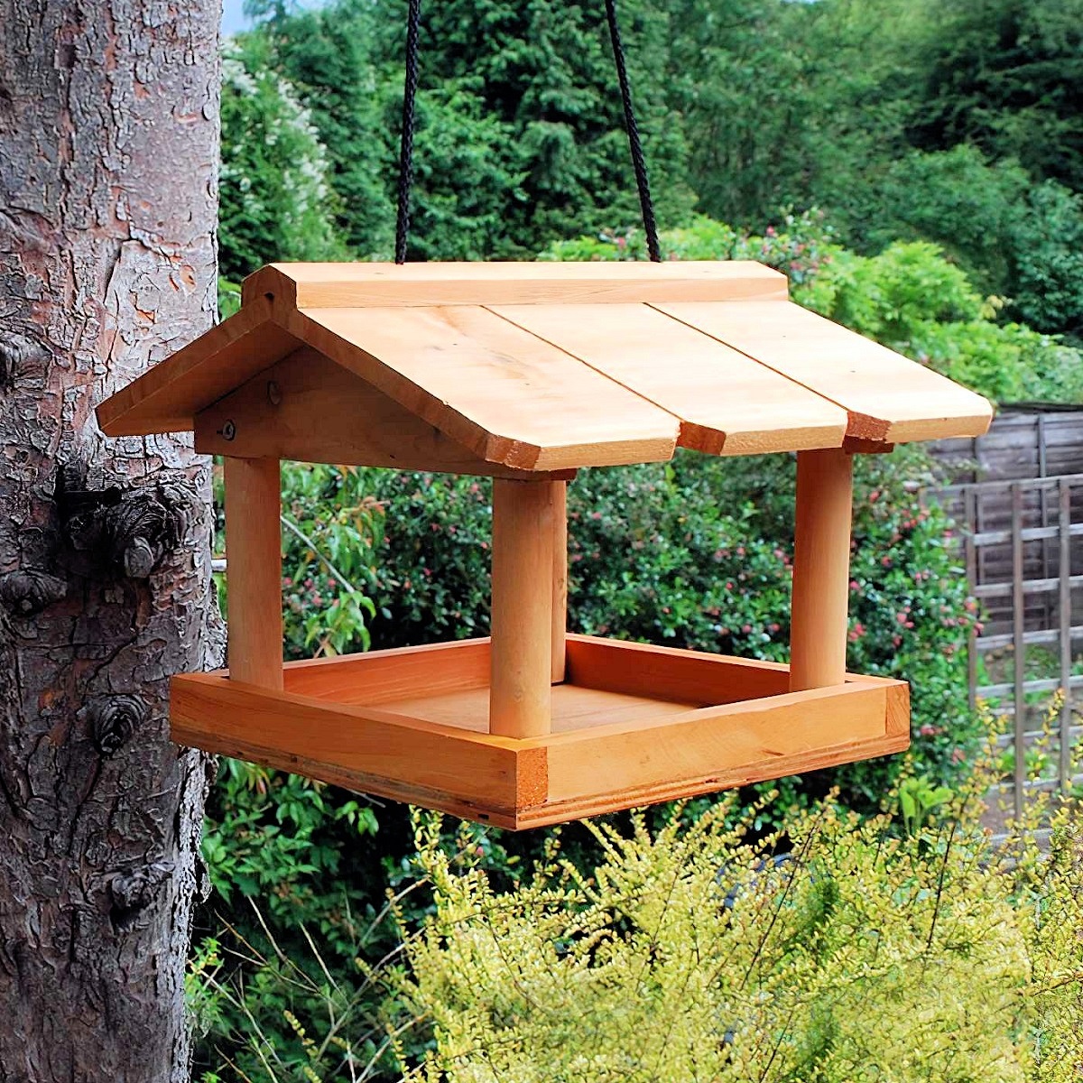 Hanging wooden bird feeder with a roof, suspended from a tree in a garden setting.