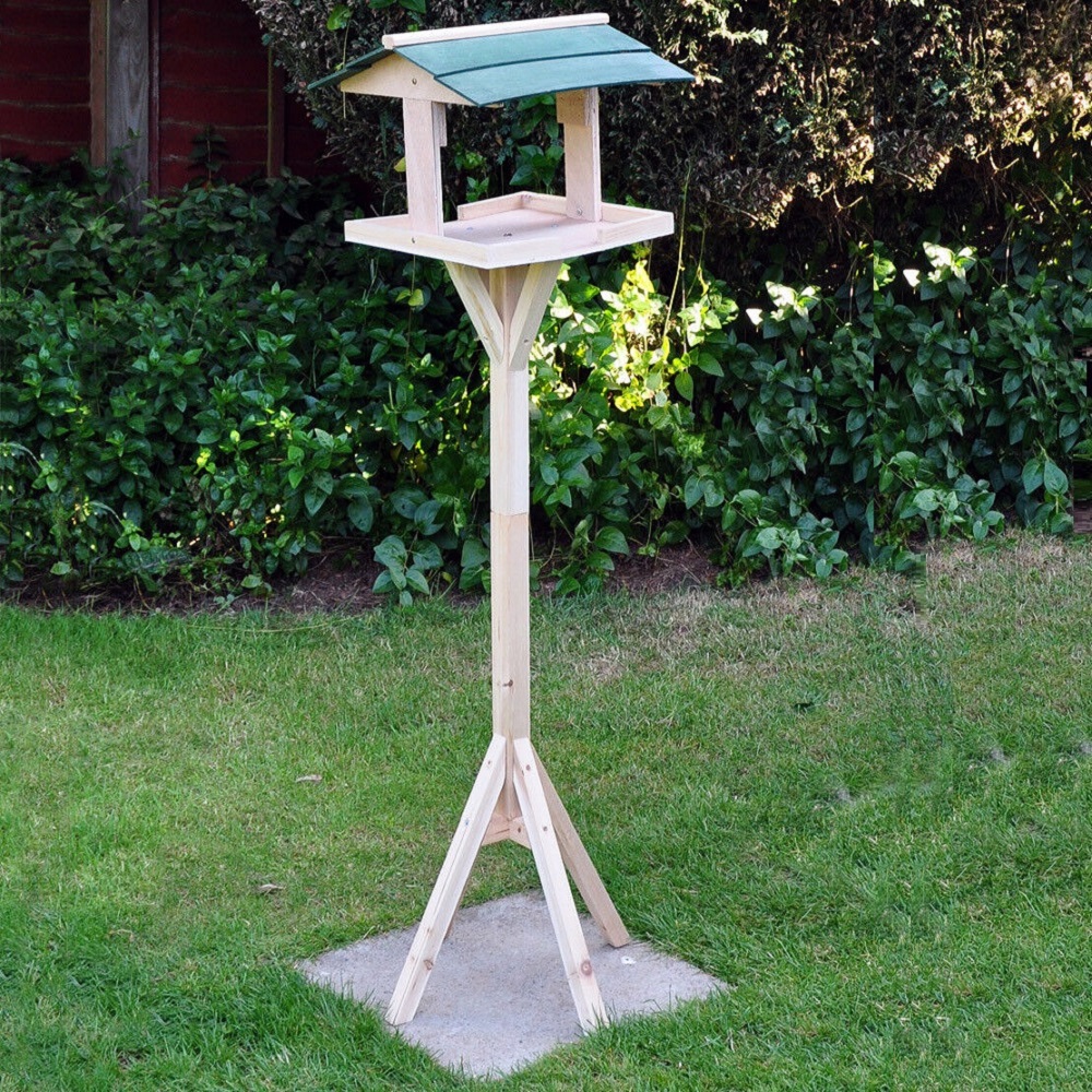 Wooden garden bird table with a green roof, standing on grass with bushes in the background.