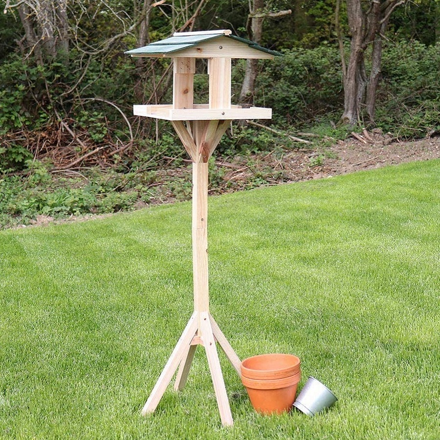 Wooden garden bird table with a green roof, standing on a lawn with a terracotta pot and a metal bucket nearby.