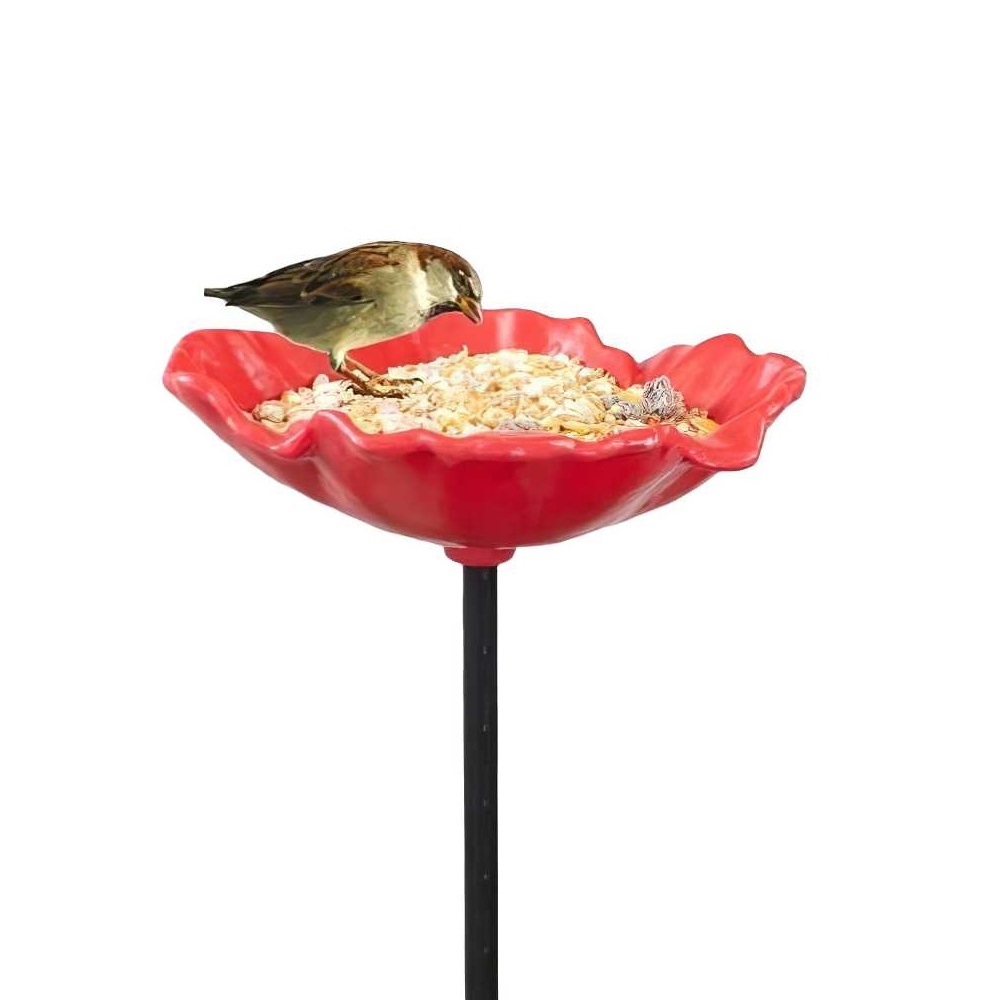 A small bird feeding from a red poppy-shaped bird feeder filled with seeds.