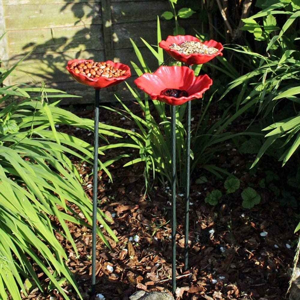 Three red poppy-shaped bird feeders filled with seeds and nuts, set in a garden with green foliage.