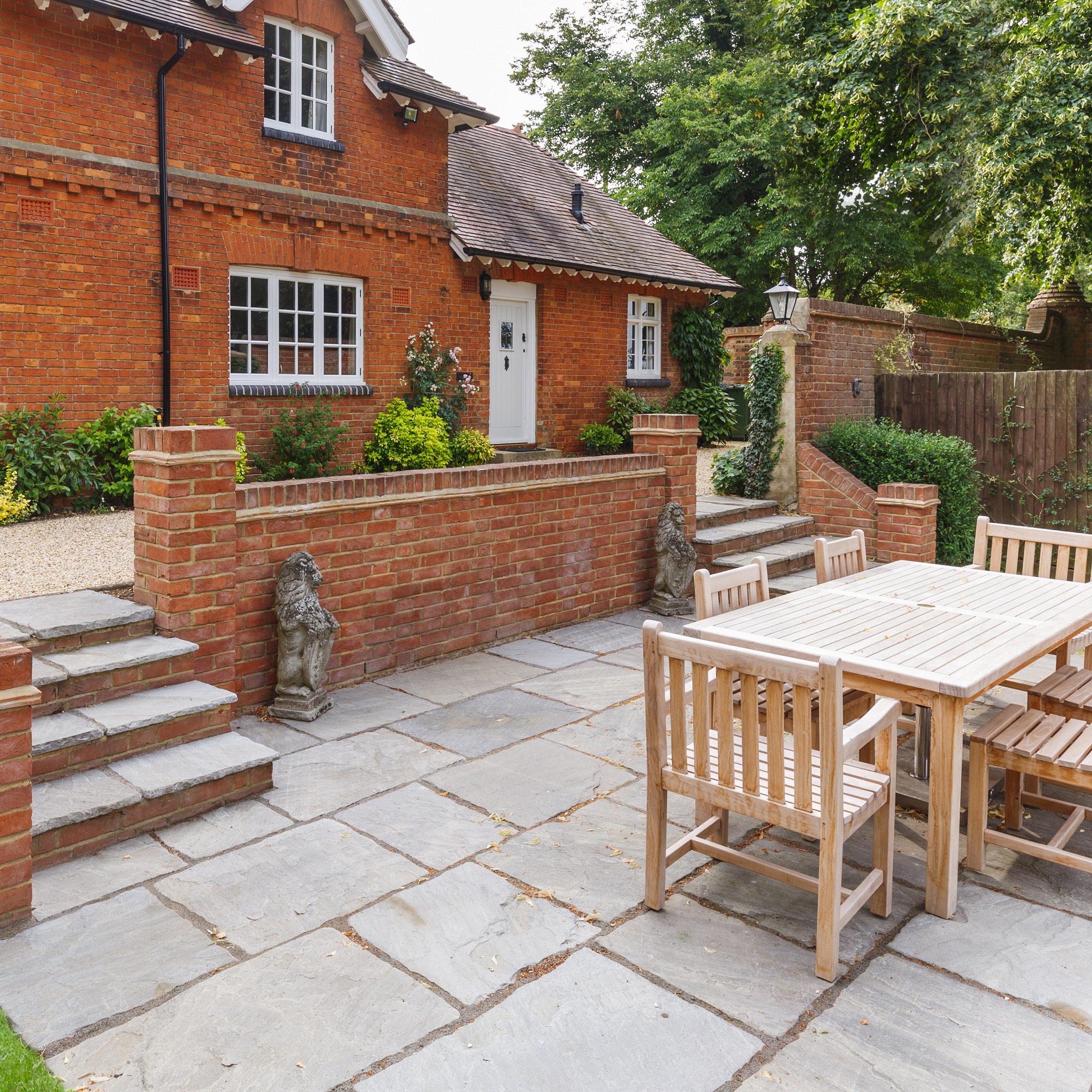 A patio area with Indian sandstone paving and wooden garden furniture in front of a red brick house.