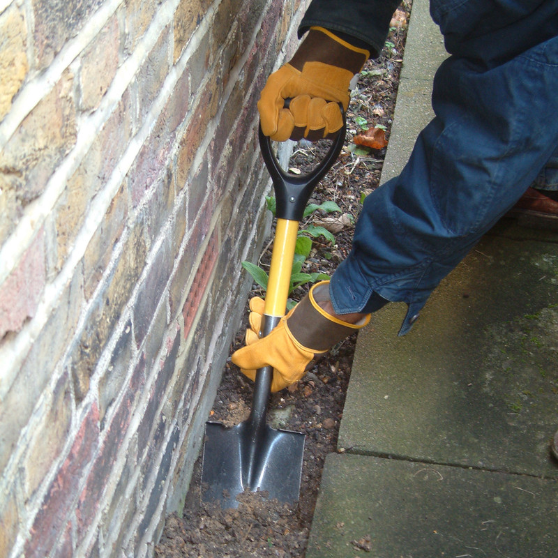 Person using a Roughneck micro square head shovel with short handle in a narrow space beside a brick wall.