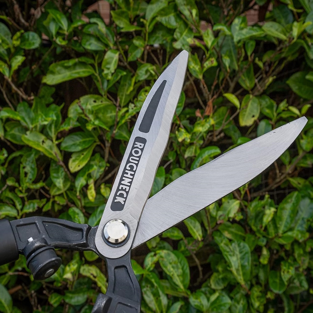 Close-up of Roughneck XT PRO Heavy Duty Wavy Blade Hedge and Grass Shears in front of green foliage.