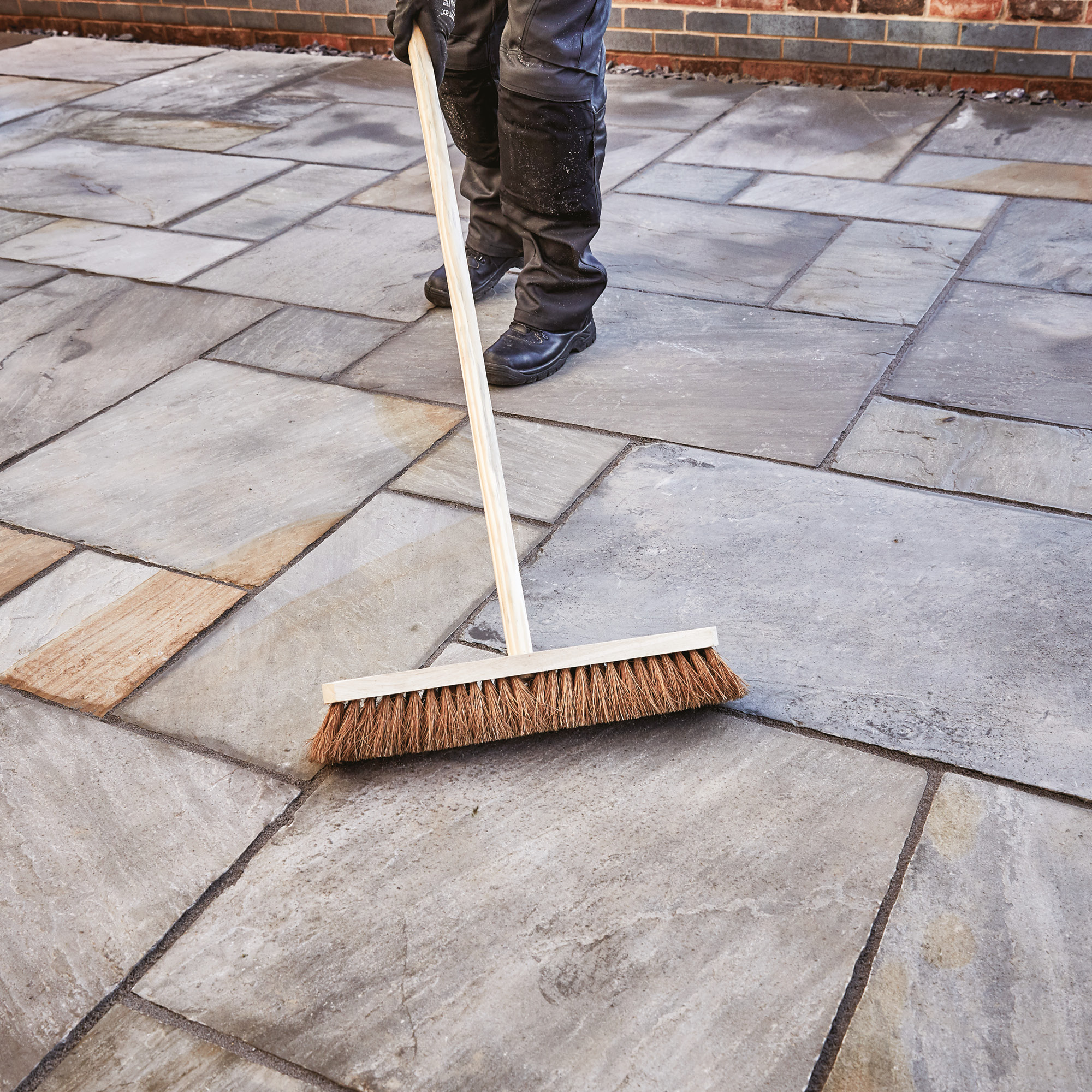 Worker sweeping jointing compound into gaps between stone patio slabs using a stiff-bristled broom