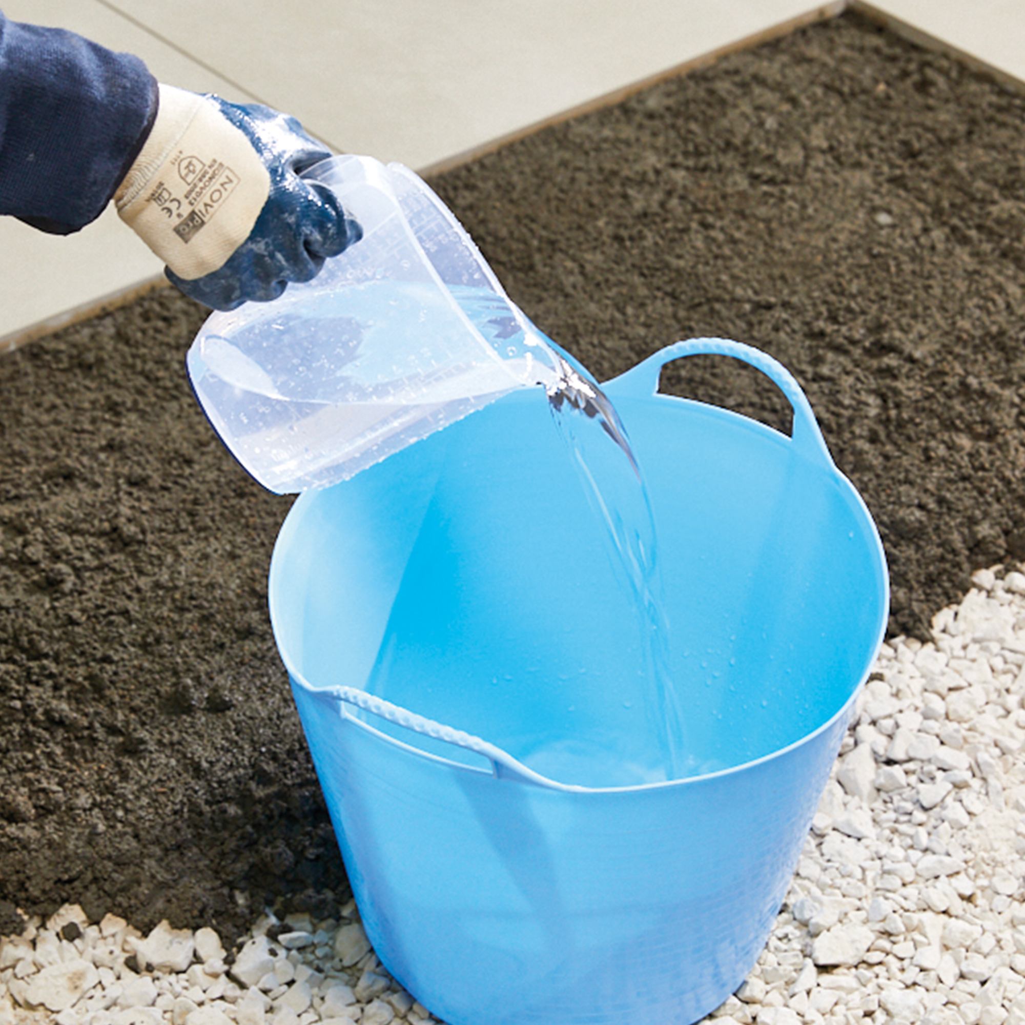 Person pouring water into a blue bucket on a gravel surface.