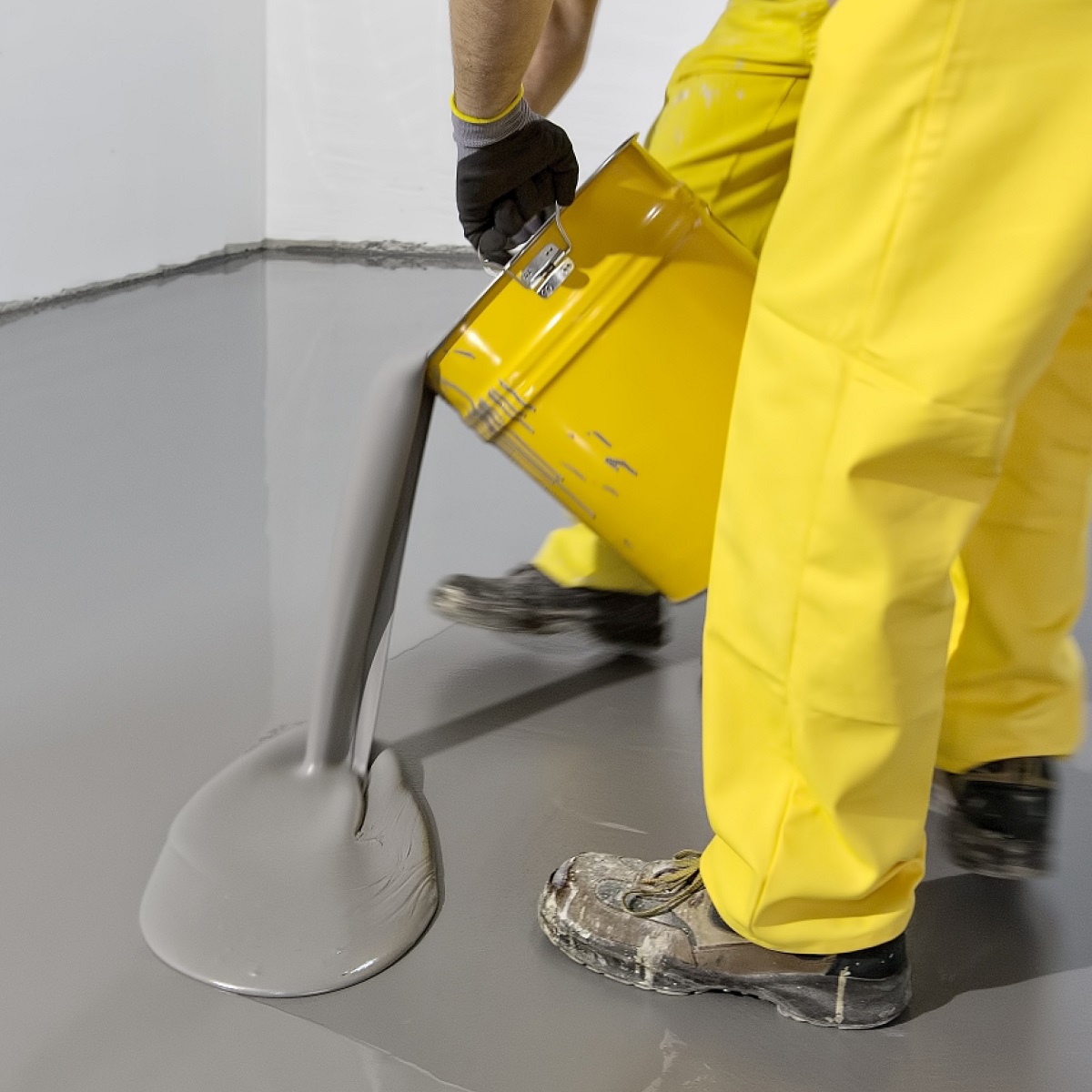 Person pouring grey self-levelling compound on floor from yellow container.