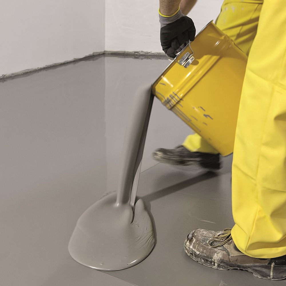 Person pouring grey self-levelling compound onto a floor from a yellow container.