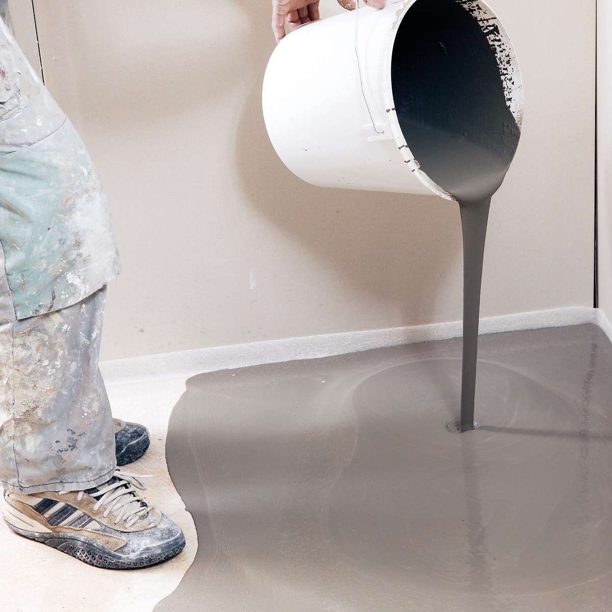 Person pouring grey self-levelling compound onto a floor from a white bucket.