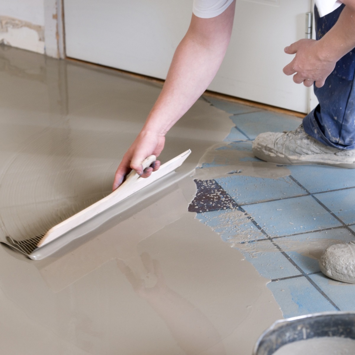 Person applying self-levelling compound on a tiled floor using a trowel.
