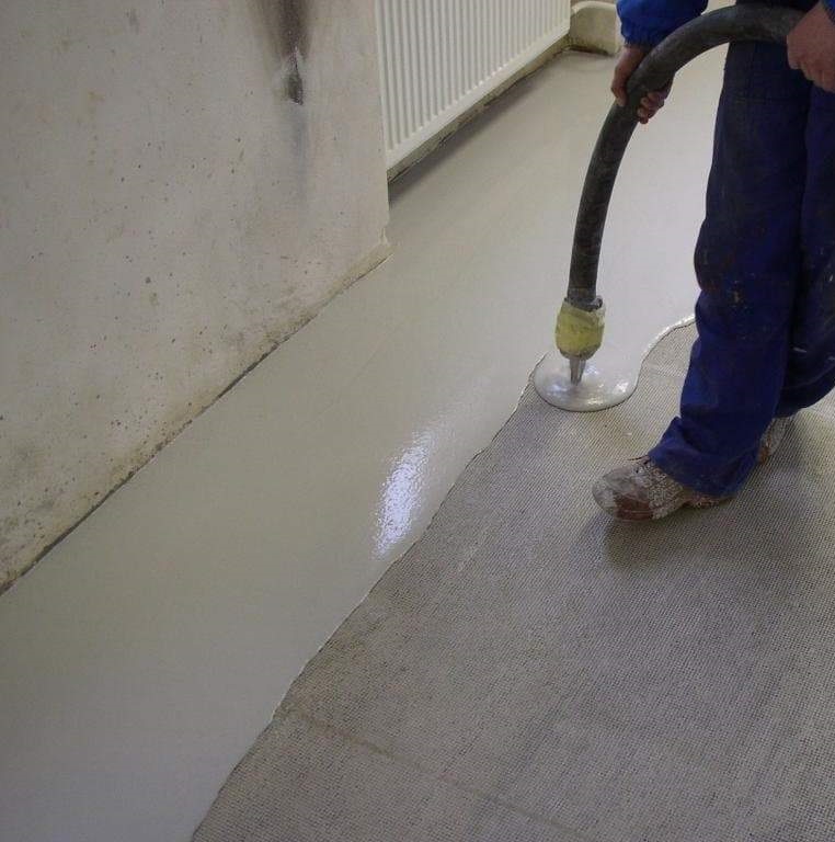 Person applying self-levelling concrete floor compound with a hose in a room.