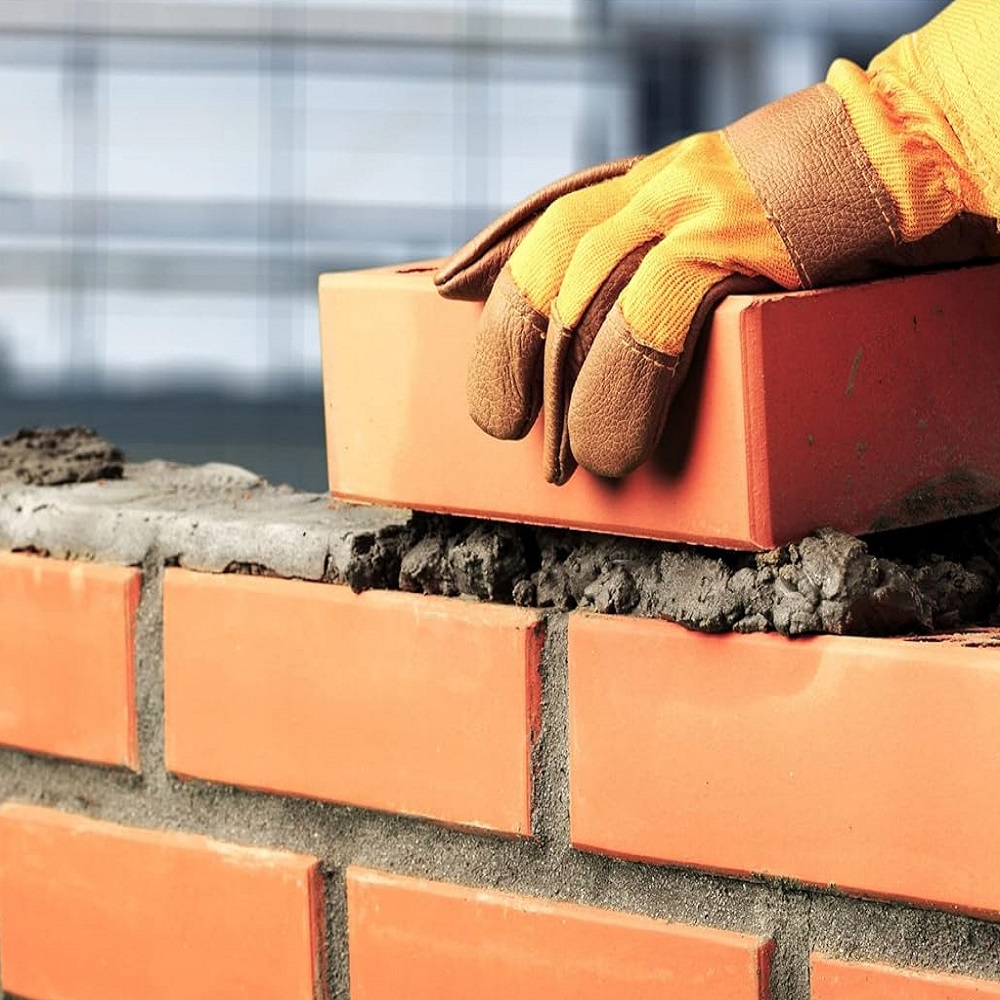A gloved hand placing a brick on a wall with mortar.