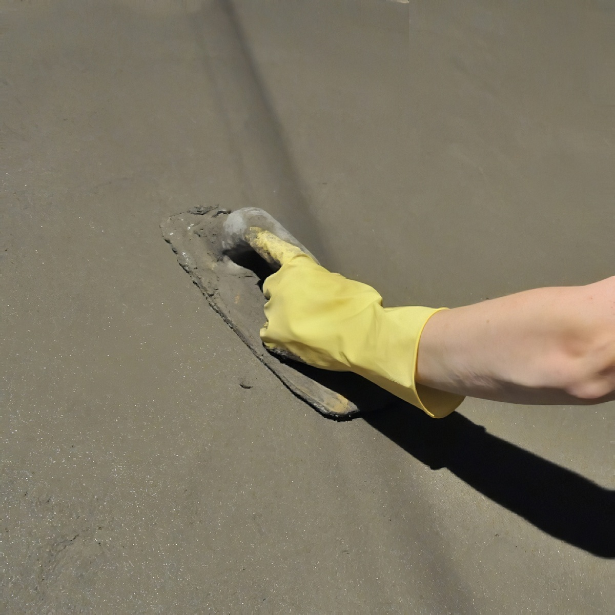 Person smoothing concrete with a trowel wearing yellow gloves and a white helmet.