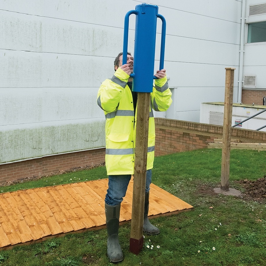 Person using a blue Silverline Pro Steel Rammer Tube Driver to install a wooden fence post in a garden area.