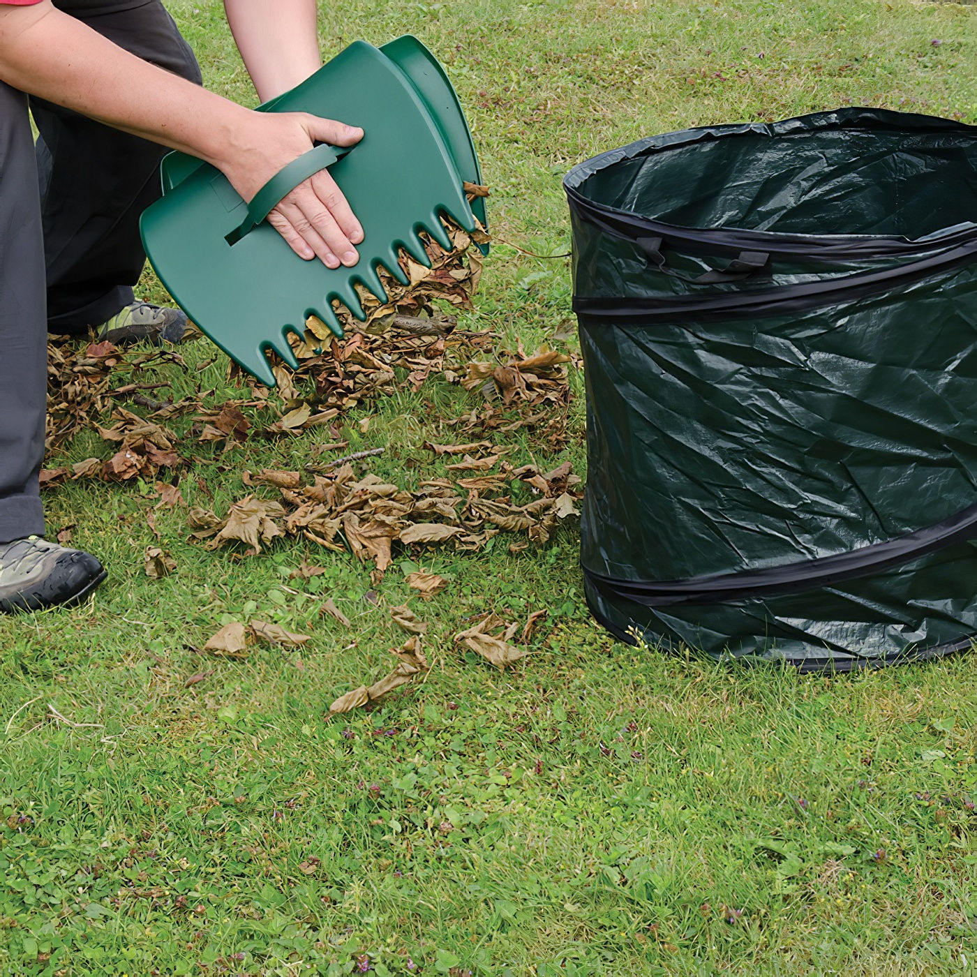 Person using green leaf collector scoops to gather leaves into a garden bag on grass.