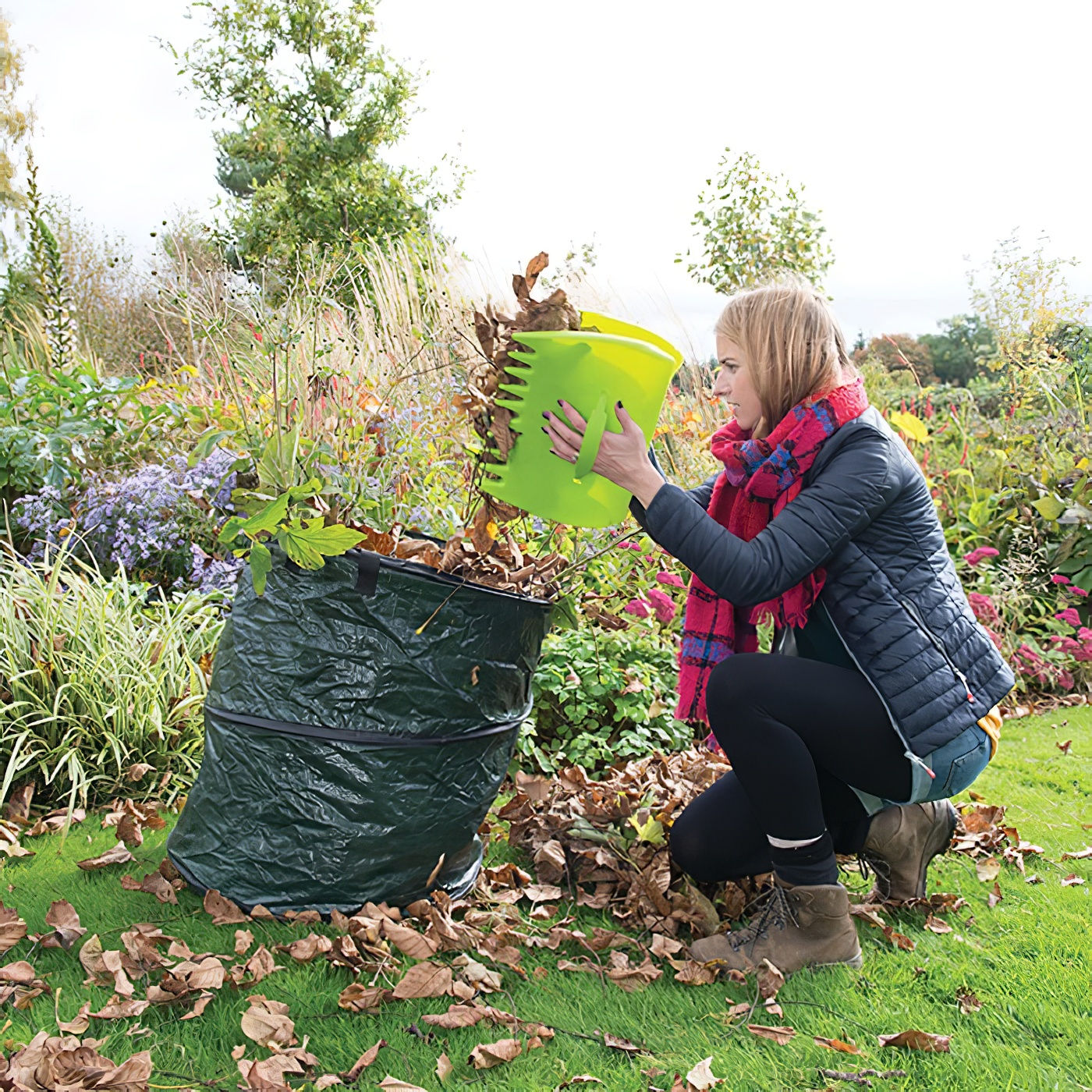 Person using green scoops to collect leaves in a garden, placing them into a large green bag.