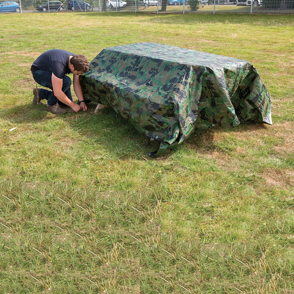 Person securing a green camouflage tarpaulin sheet over an object on grass.