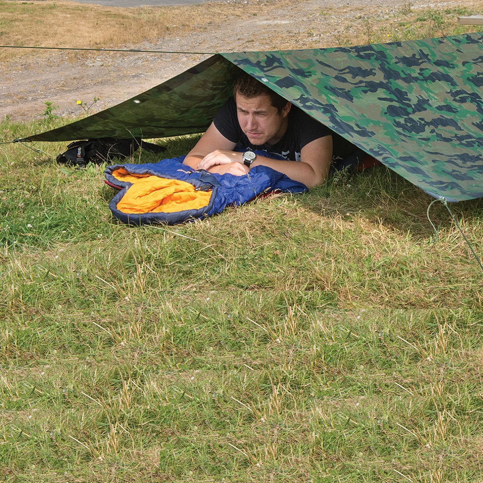 Person lying under a green camouflage tarpaulin sheet on grass.