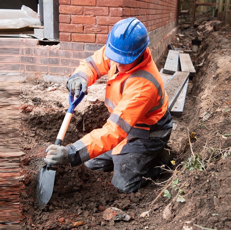 Worker using a stubby short round head shovel for digging in a narrow trench.